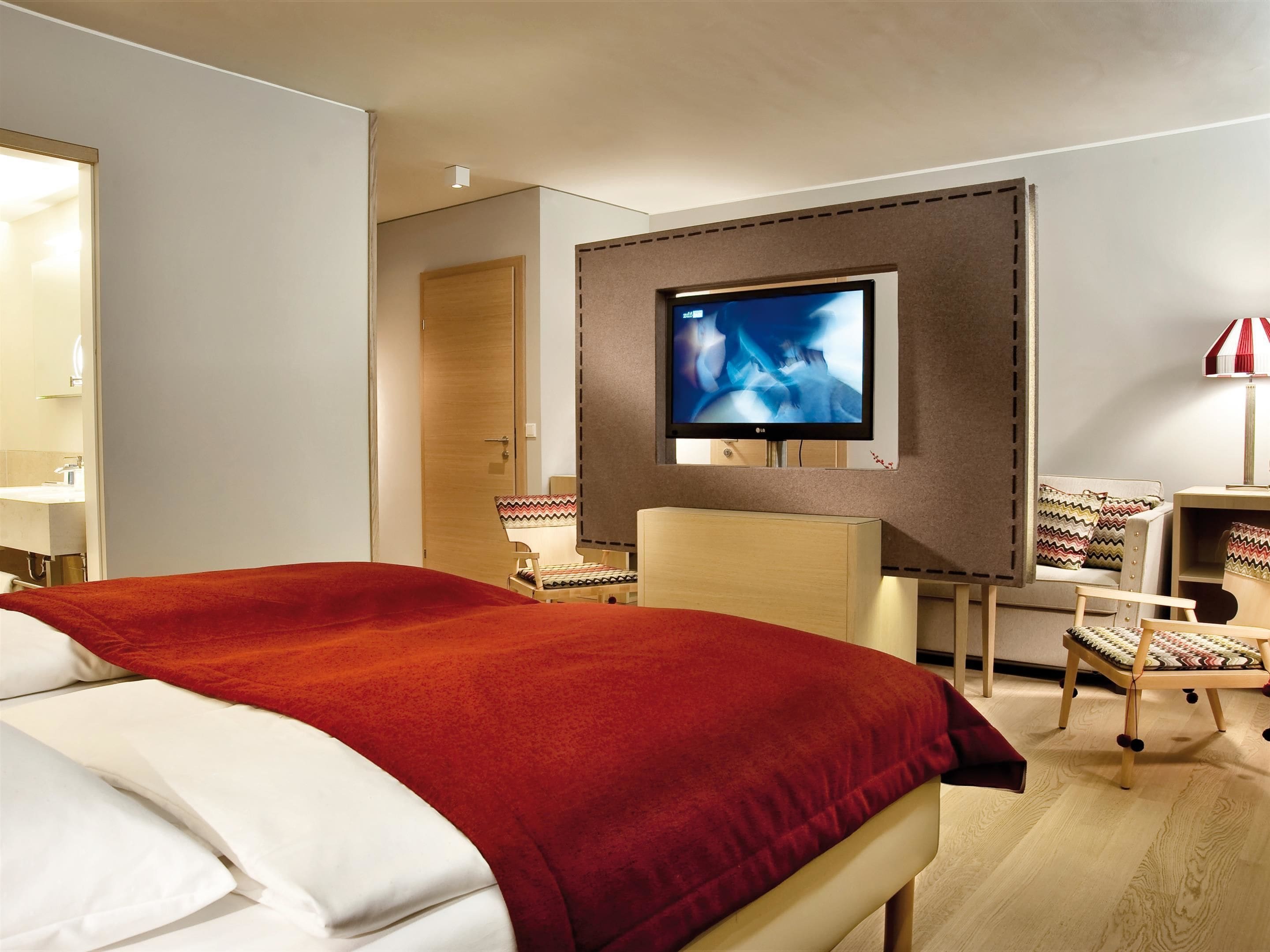 Een hotelkamer met een groot bed, een aan de muur bevestigde televisie, twee stoelen, een bureau en een lamp.