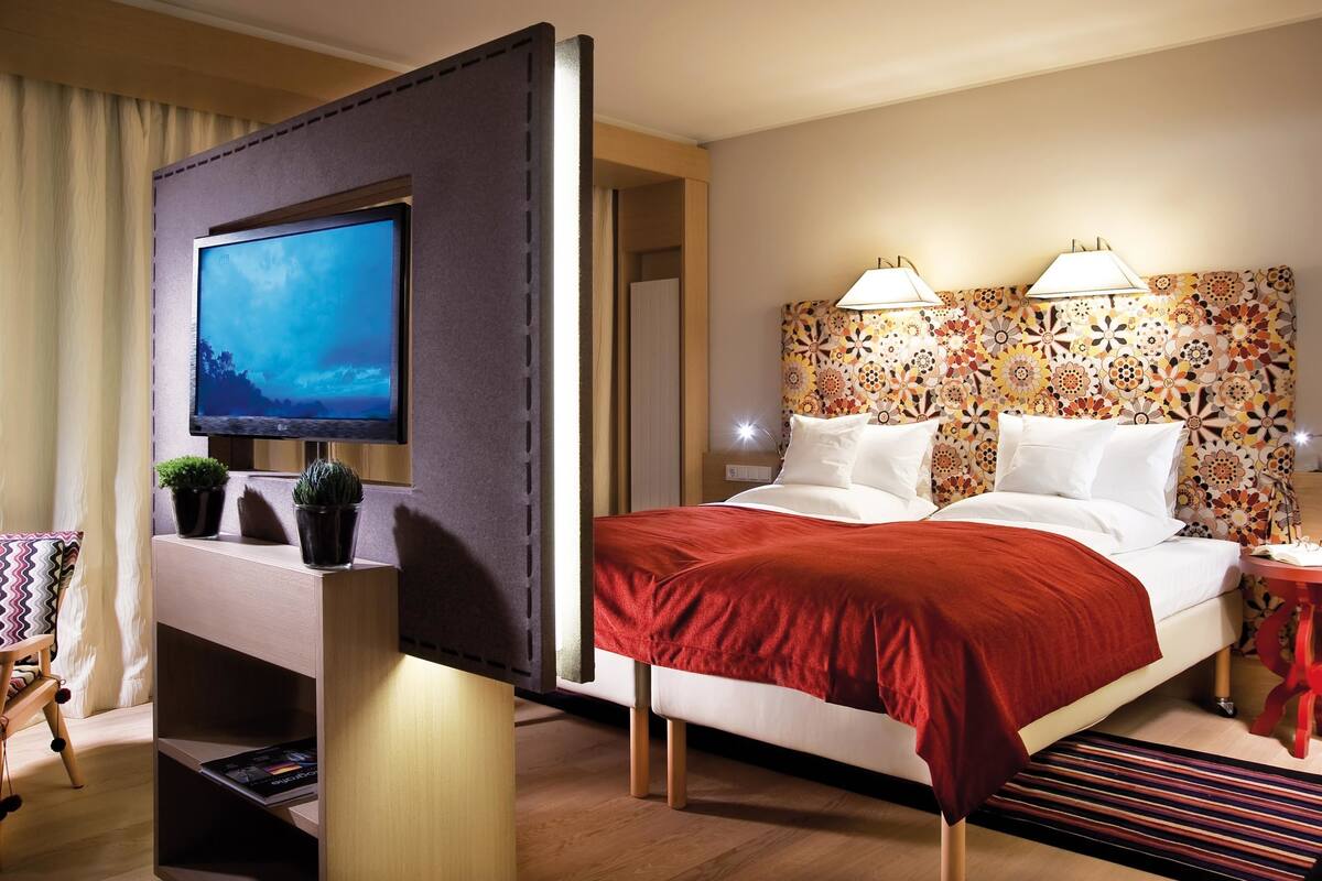 A hotel room with a bed, a TV mounted on a partition, a patterned headboard, and a small bedside table with a lamp.