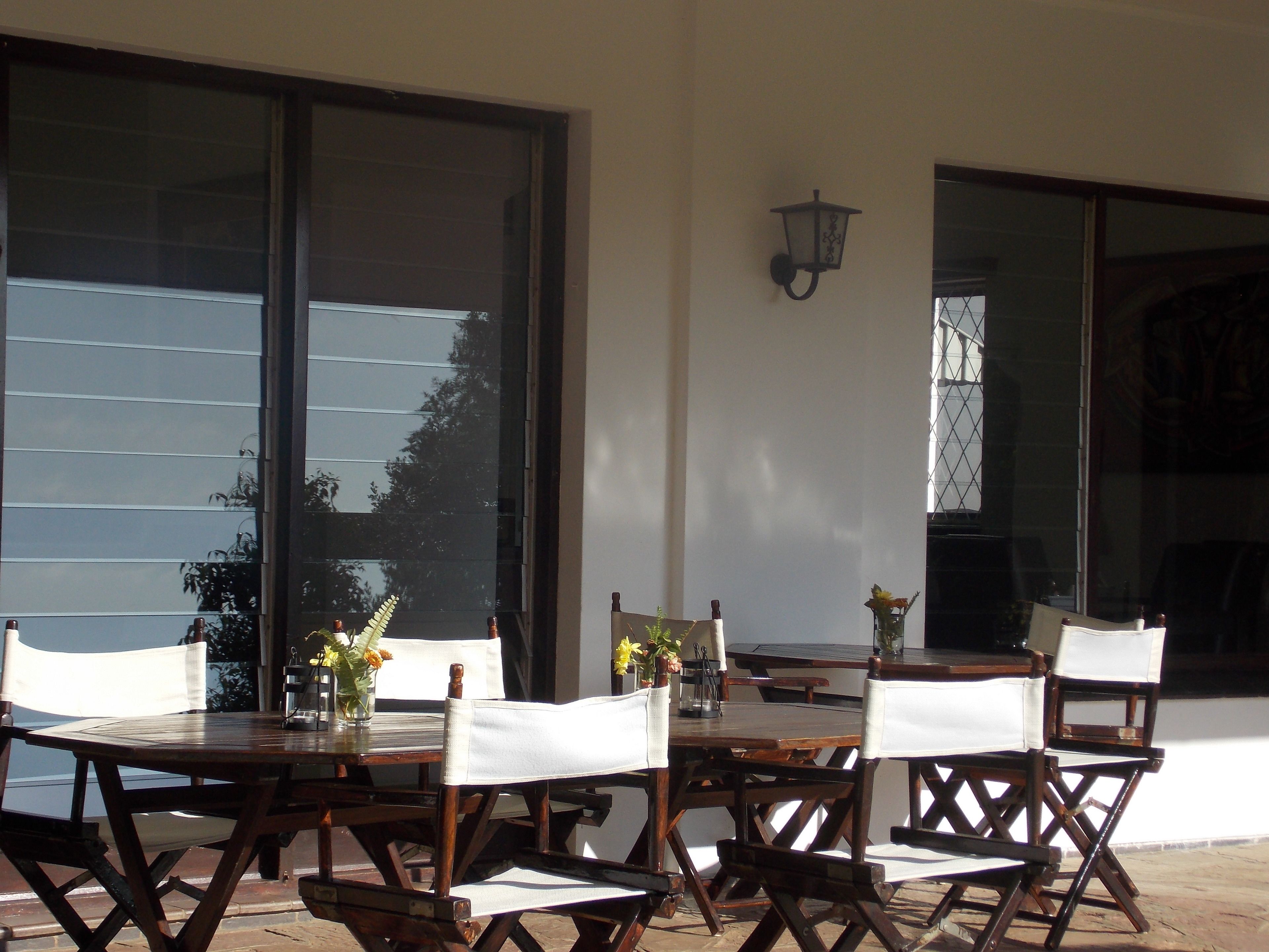 Un patio exterior con una mesa y sillas de madera, un aplique de pared y ventanas amplias que reflejan el cielo y los árboles.