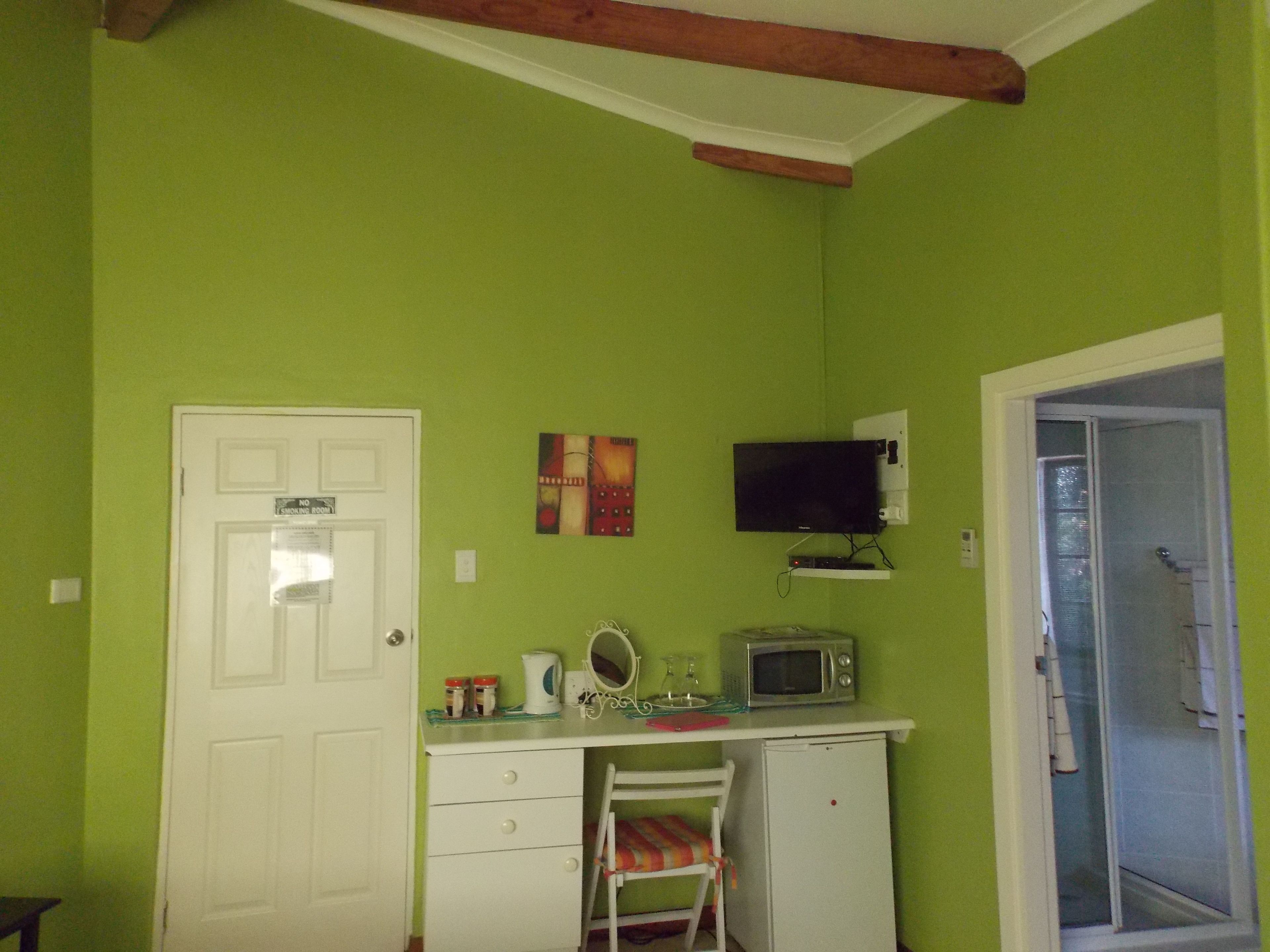 A room with a green wall, a white door, a small white desk with a microwave, a chair, and a television mounted on the wall.