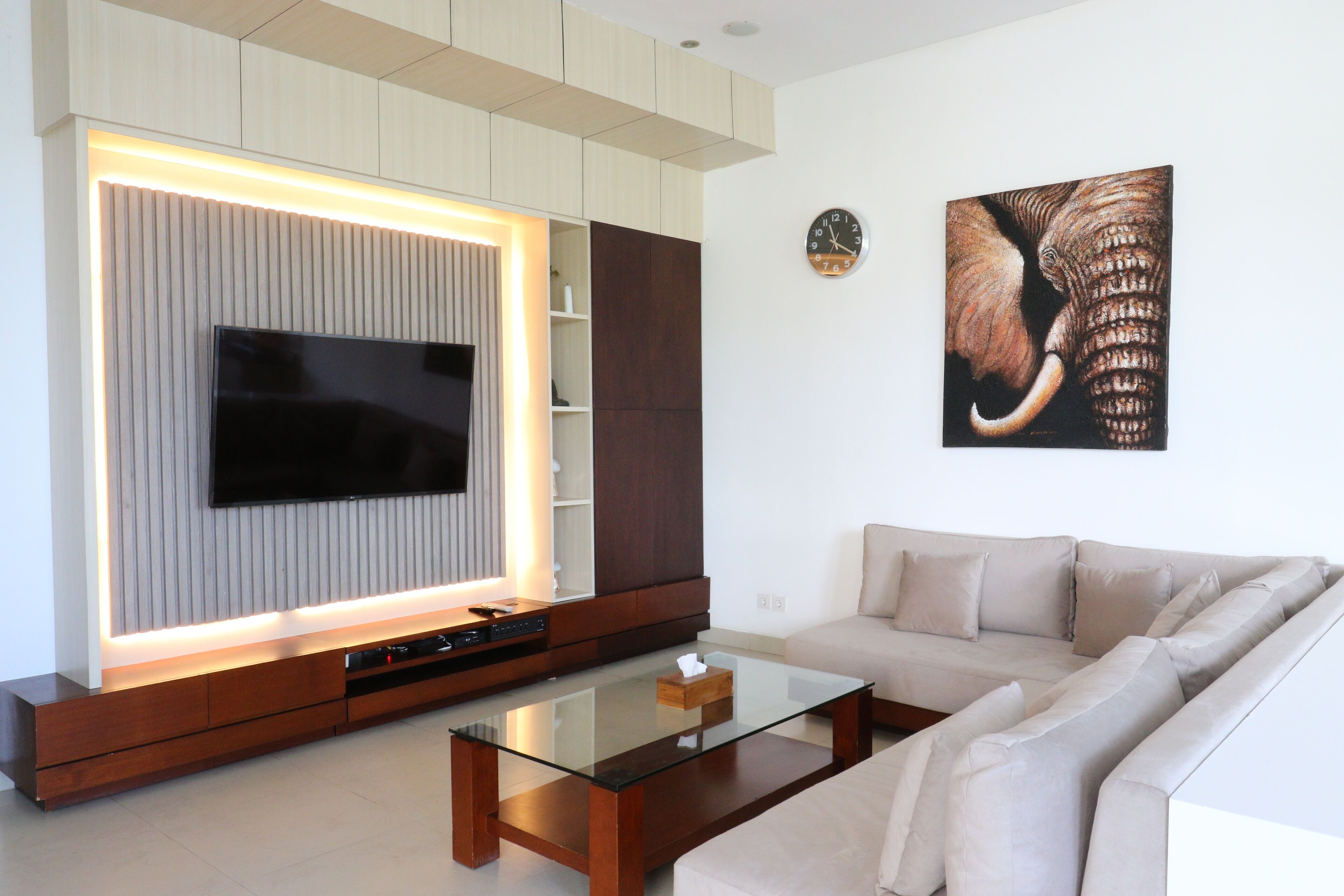 A modern living room with a flat-screen TV, a glass coffee table, a beige sofa, and a wooden entertainment unit.