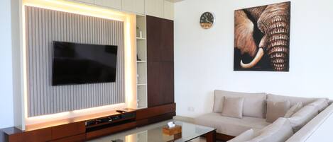 A modern living room with a flat-screen TV, a glass coffee table, a beige sofa, and a wooden entertainment unit.