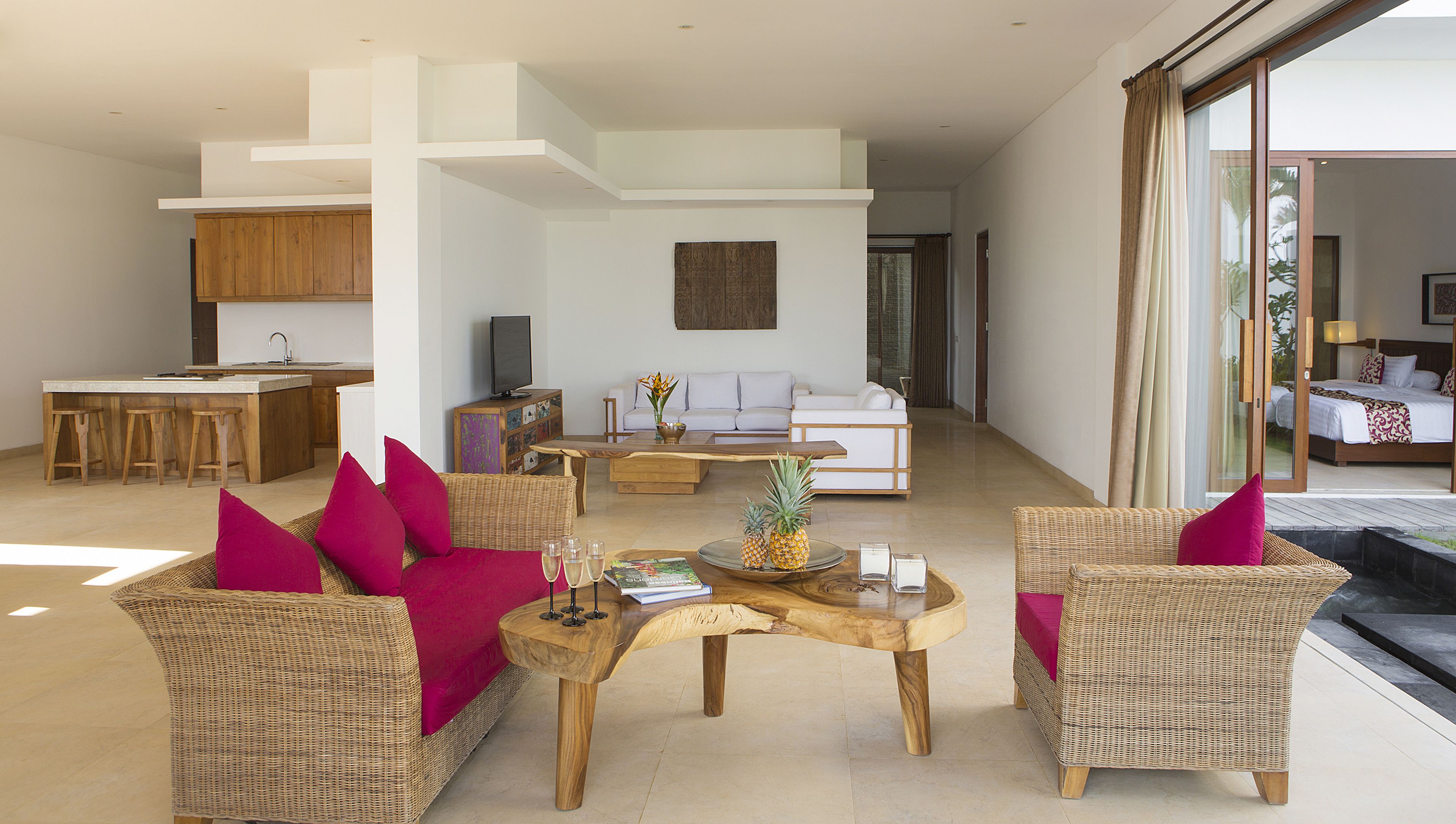 A spacious living area with wicker furniture, a coffee table, and a sofa with bright red cushions. There is a kitchen area with wooden cabinets and a dining table with bar stools.
