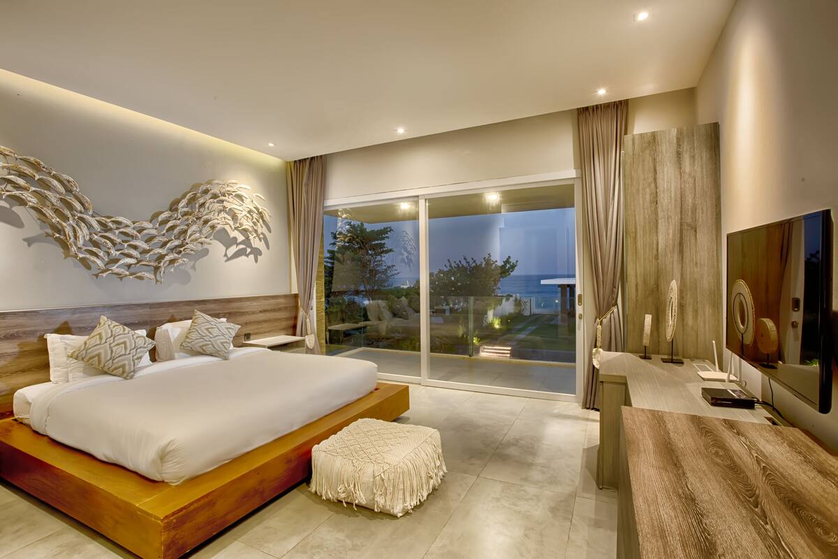 A modern bedroom with a large bed, a wooden bench, a TV, and a balcony with a view.