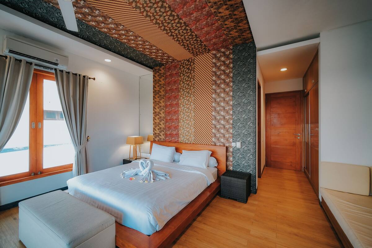 A hotel room with a large bed, a wooden floor, a patterned wall, and a window with curtains.