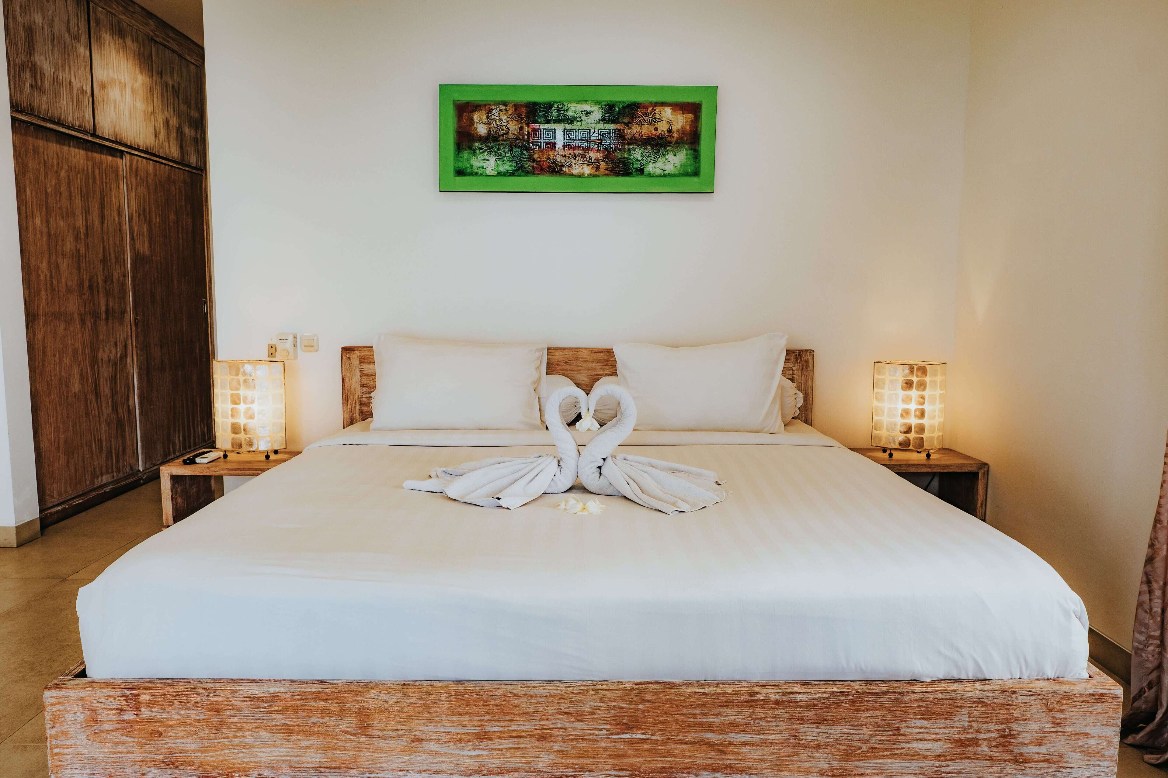 Una cama bien hecha con una decoración de toalla con forma de cisne, dos lámparas de noche y una obra de arte con marco verde en la pared.