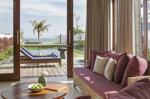 Una sala moderna con vista al océano, un sillón, un sofá con cojines y una mesa con un frutero y un libro.