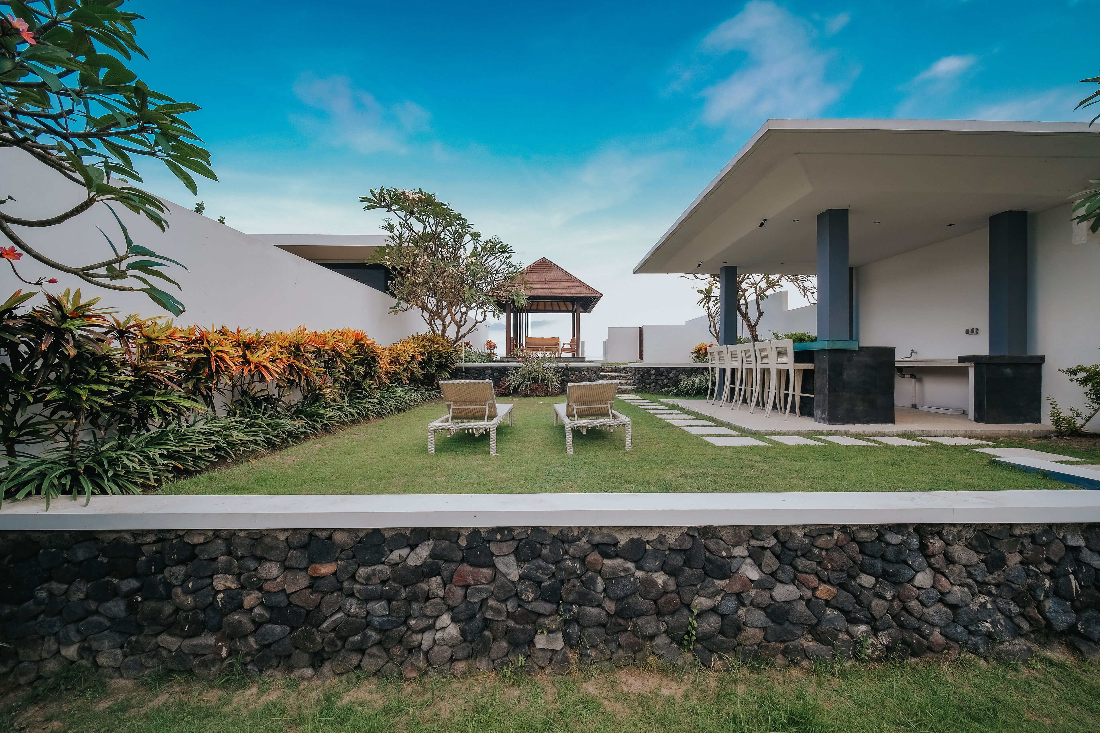 A modern outdoor area with a stone wall, two lounge chairs, a wooden gazebo, and a poolside area with a table and chairs.