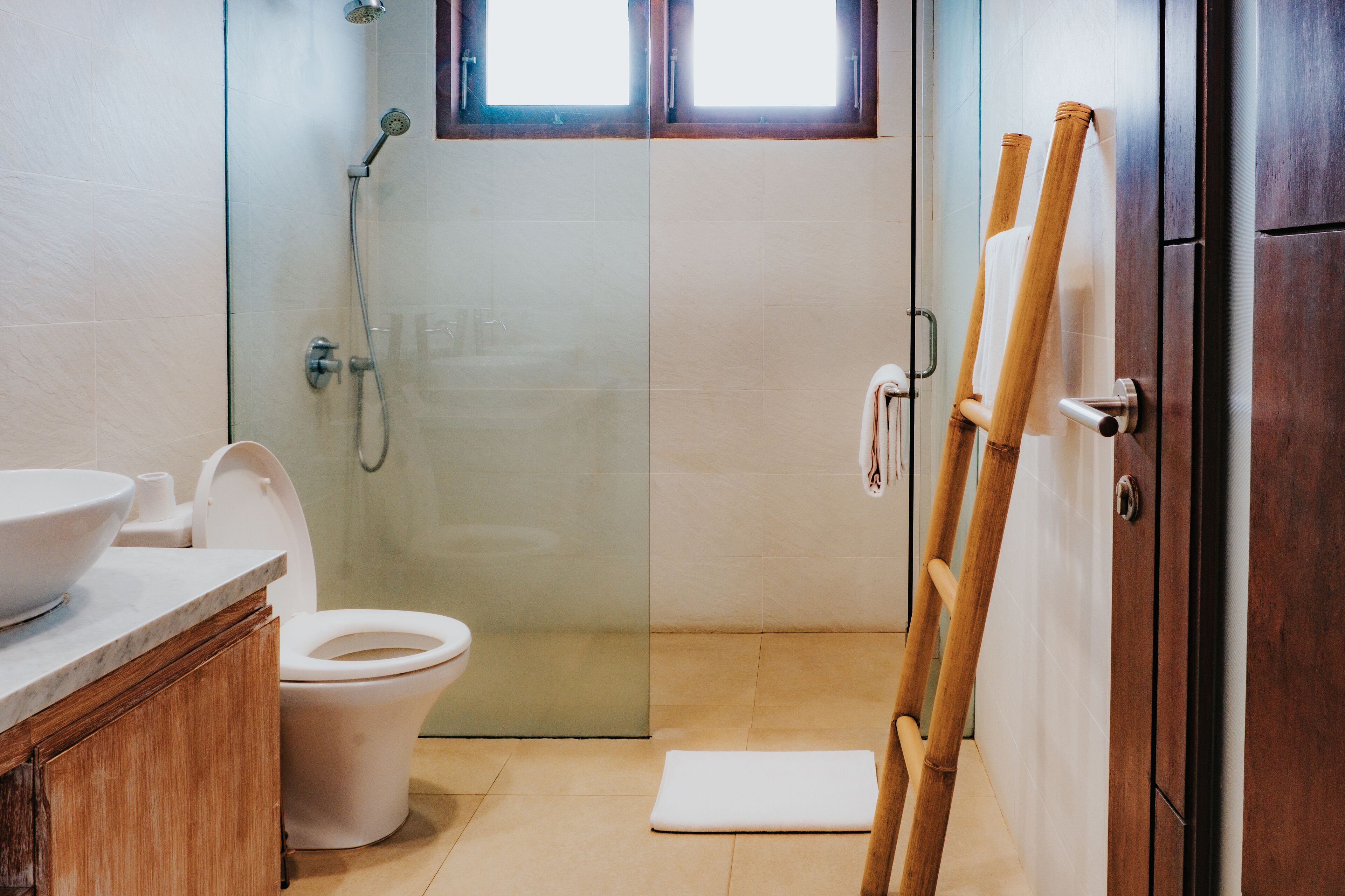 A bathroom with a glass shower enclosure, a toilet, a sink, and a wooden ladder leaning against a cabinet.