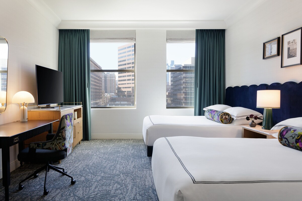 A hotel room with a desk, two beds, a TV, and a city view.