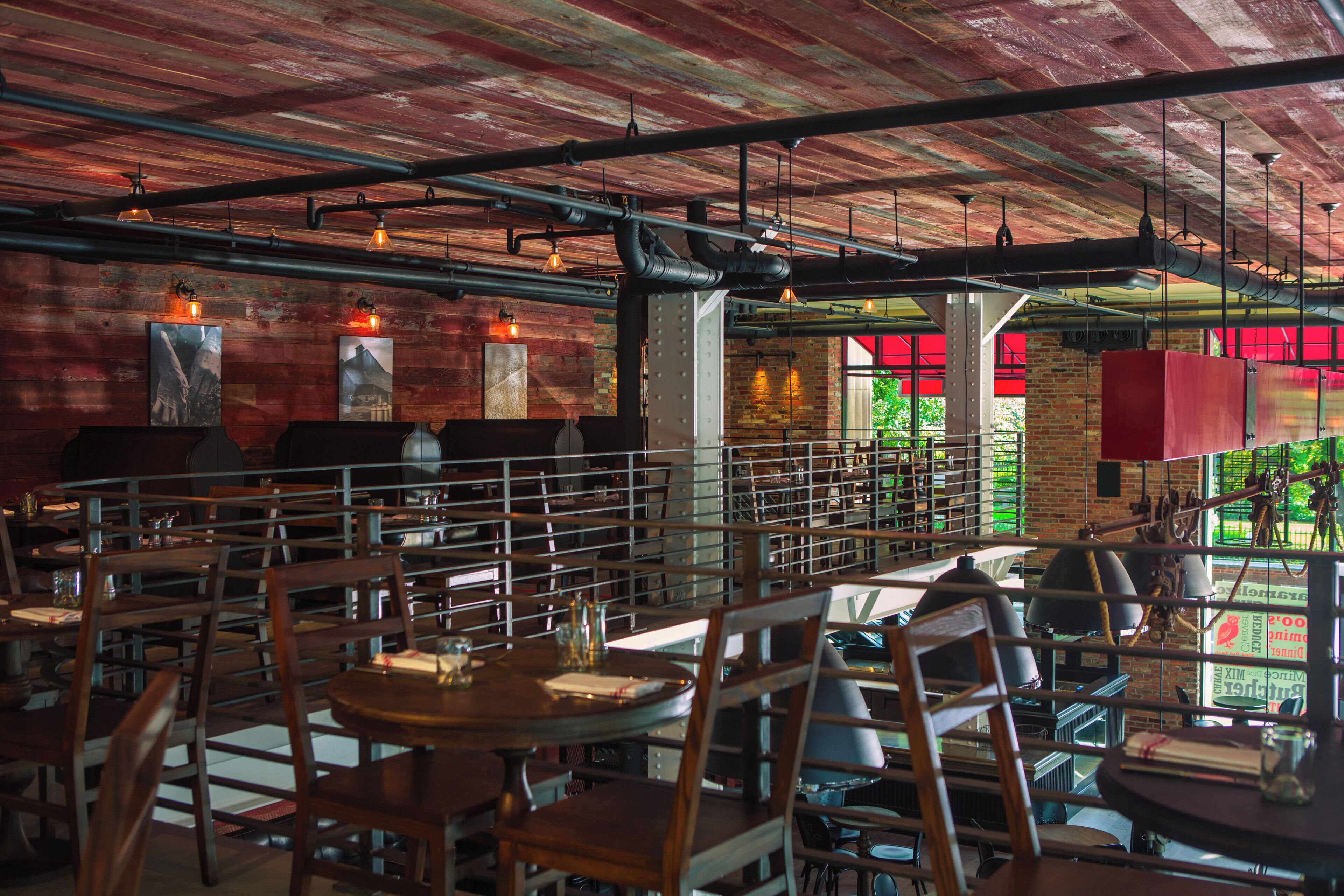 An interior with high ceilings, exposed pipes, and a mix of brick and wooden walls. Tables and chairs are arranged for dining.