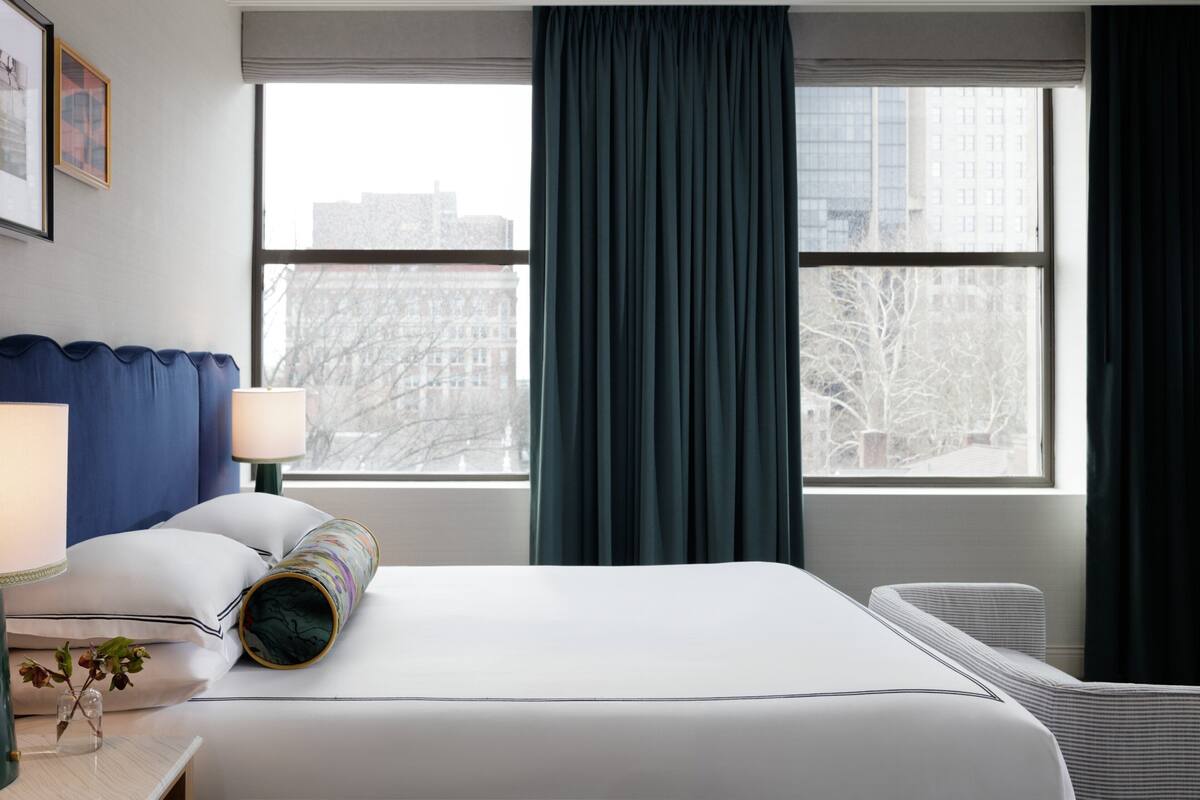 A hotel room with a bed, a nightstand with a lamp, a window with curtains, and a radiator.