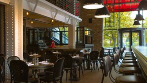 A restaurant with high ceilings, exposed beams, and a long counter with bar stools. Tables are arranged for dining, and there are hanging pendant lights.