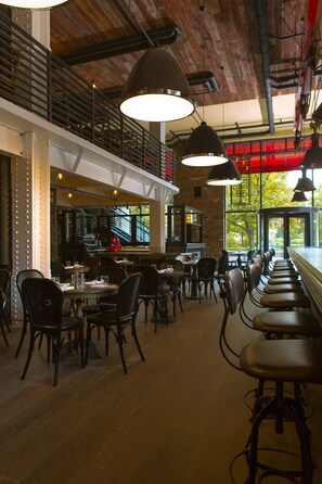 Un restaurante con techos altos, vigas a la vista y una larga barra con taburetes. Mesas dispuestas para comer y lámparas colgantes.