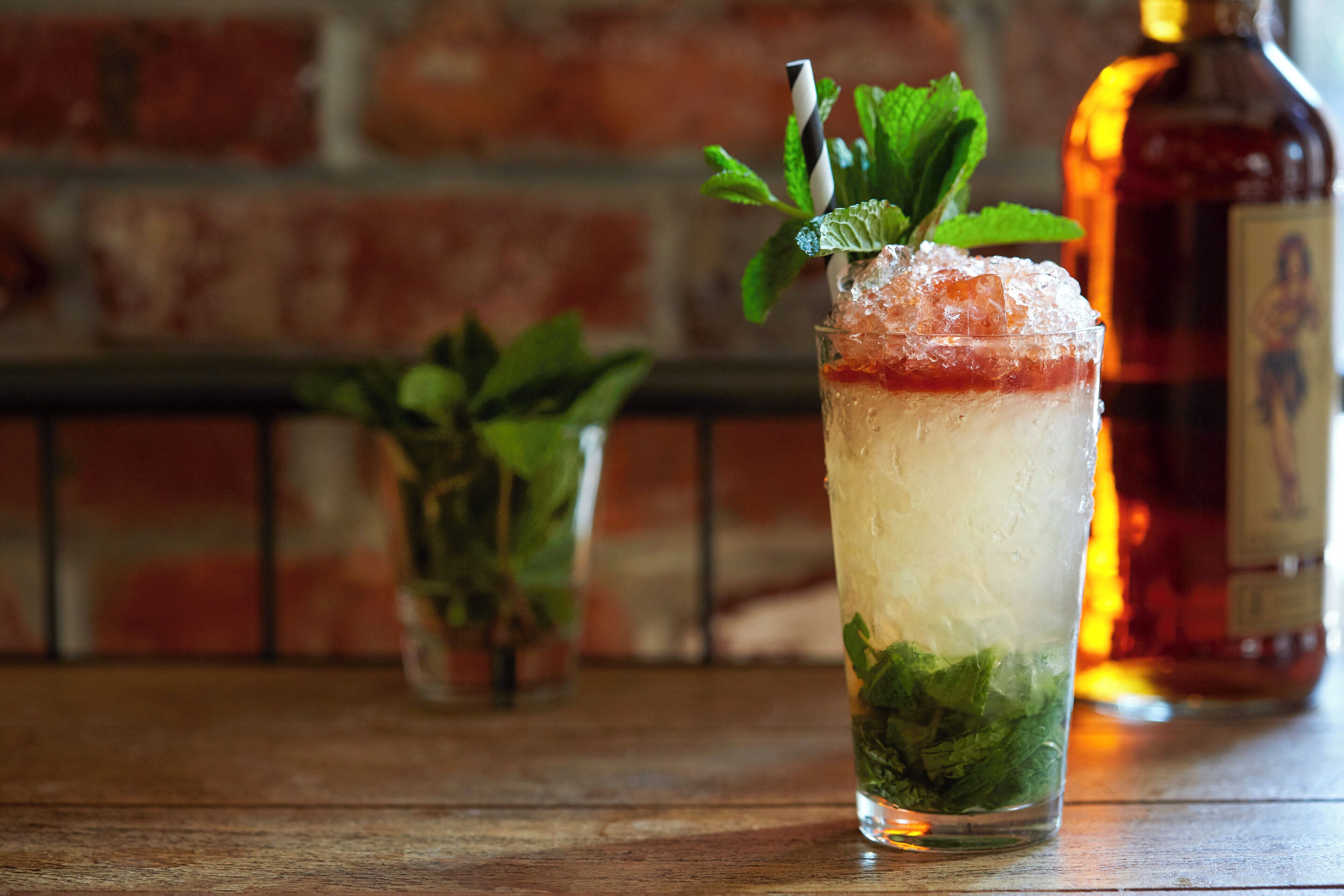 A tall glass with layered drinks, mint leaves, and a straw, placed on a wooden surface with a brick wall in the background.