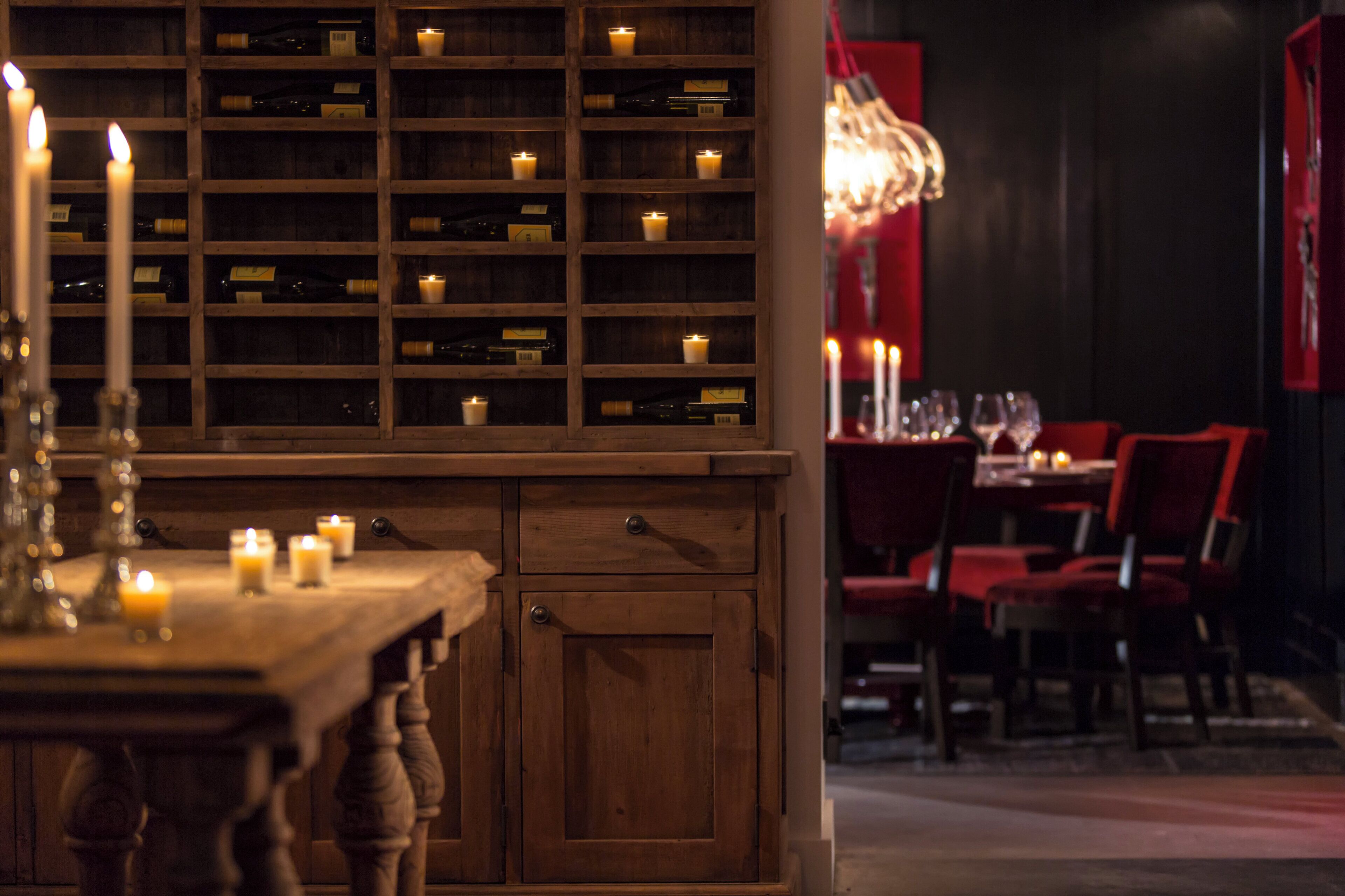 A dimly lit restaurant interior with wooden shelves displaying bottles, a table with candles, and red upholstered chairs.