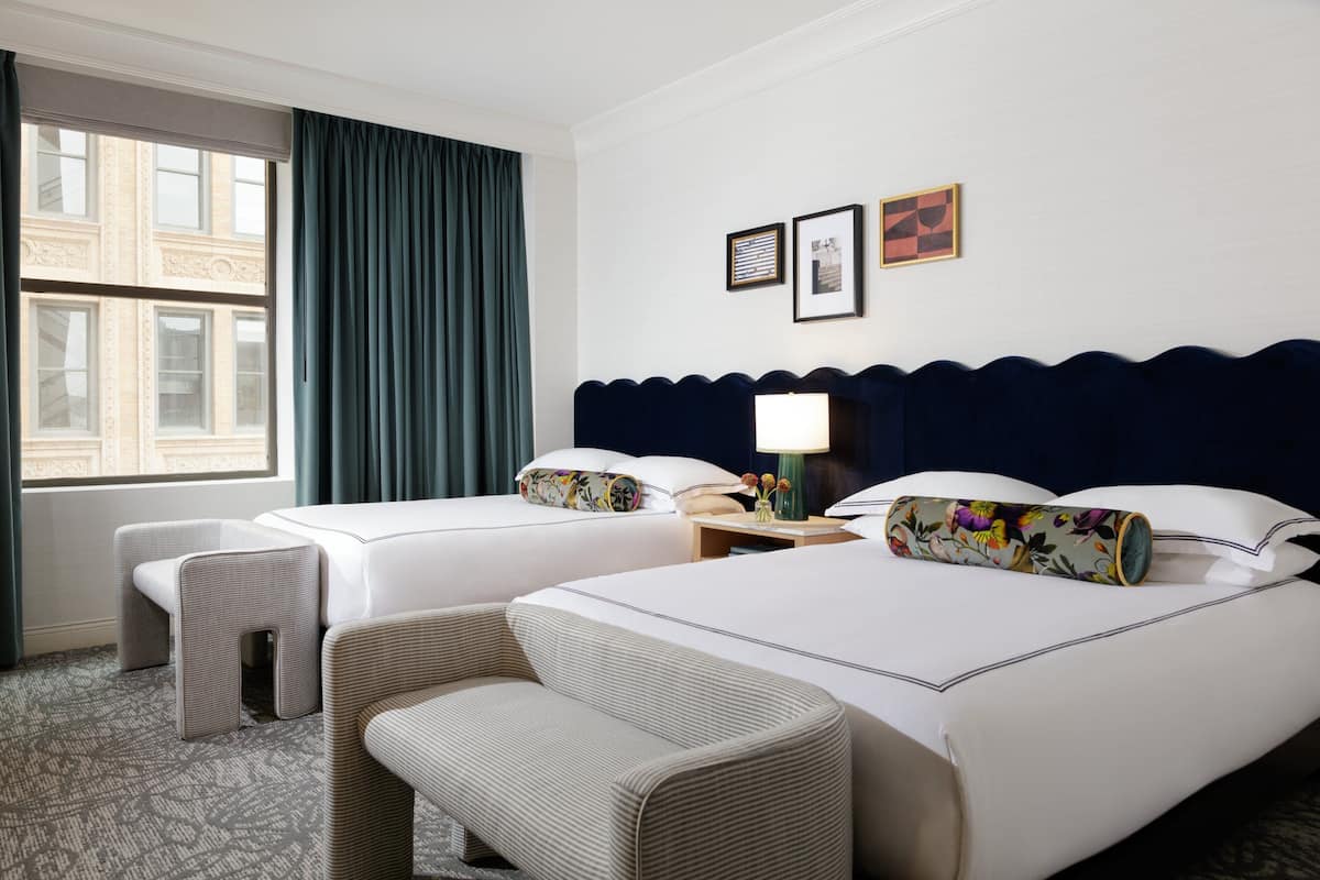 A hotel room with two beds, a nightstand, a lamp, a chair, and a window with curtains.