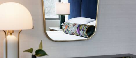 A hotel room with a bed, a bedside lamp, a small table with a vase and a notebook, and a mirror reflecting the room.
