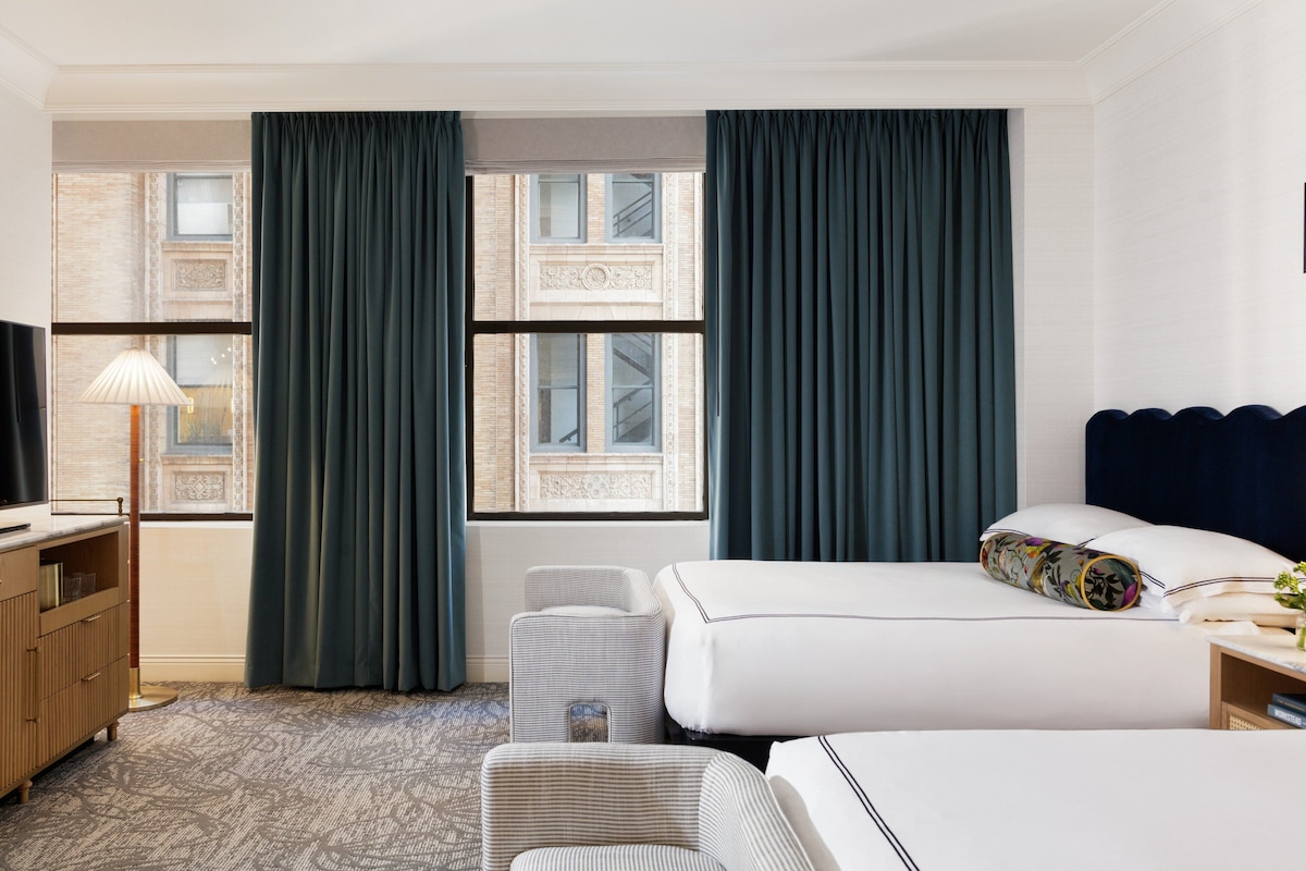 A hotel room with a bed, a desk, a lamp, and a view of the building's facade through large windows.