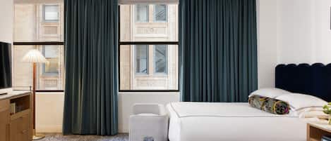 Una habitación de hotel con cama, escritorio, lámpara y vista de la fachada del edificio a través de amplias ventanas.