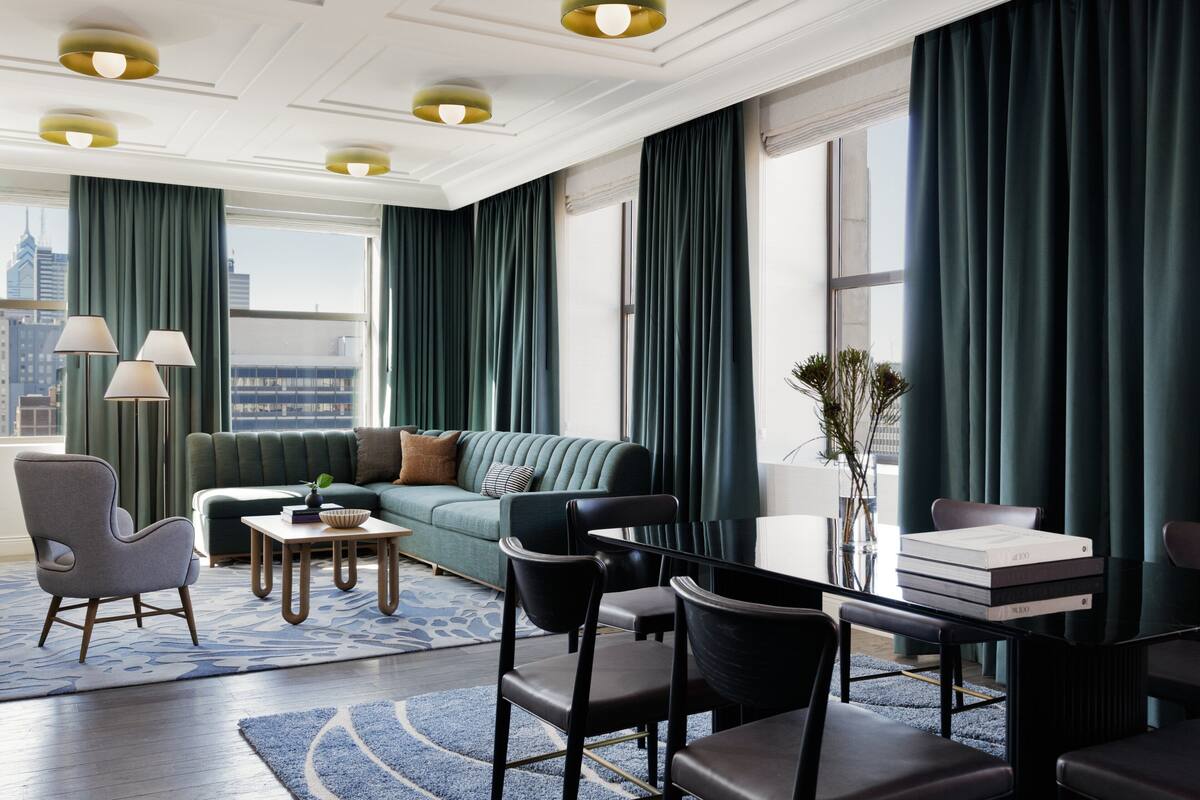 A modern hotel room with a green sofa, a dining table, chairs, a coffee table, and a view of the cityscape.