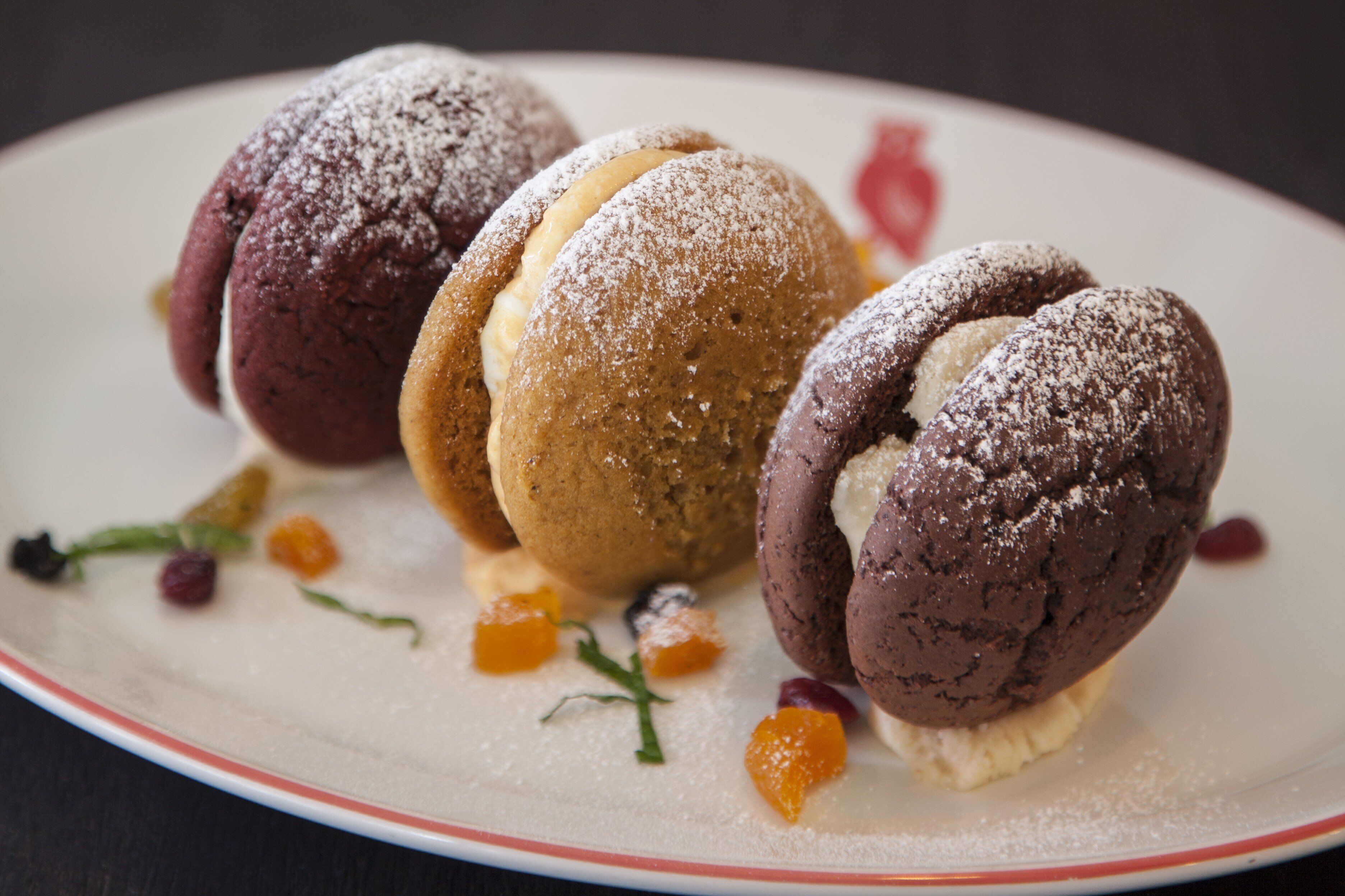 Une assiette contenant trois desserts au chocolat, chacun saupoudré de sucre glace, accompagnée de petits morceaux de fruits orange et rouge.