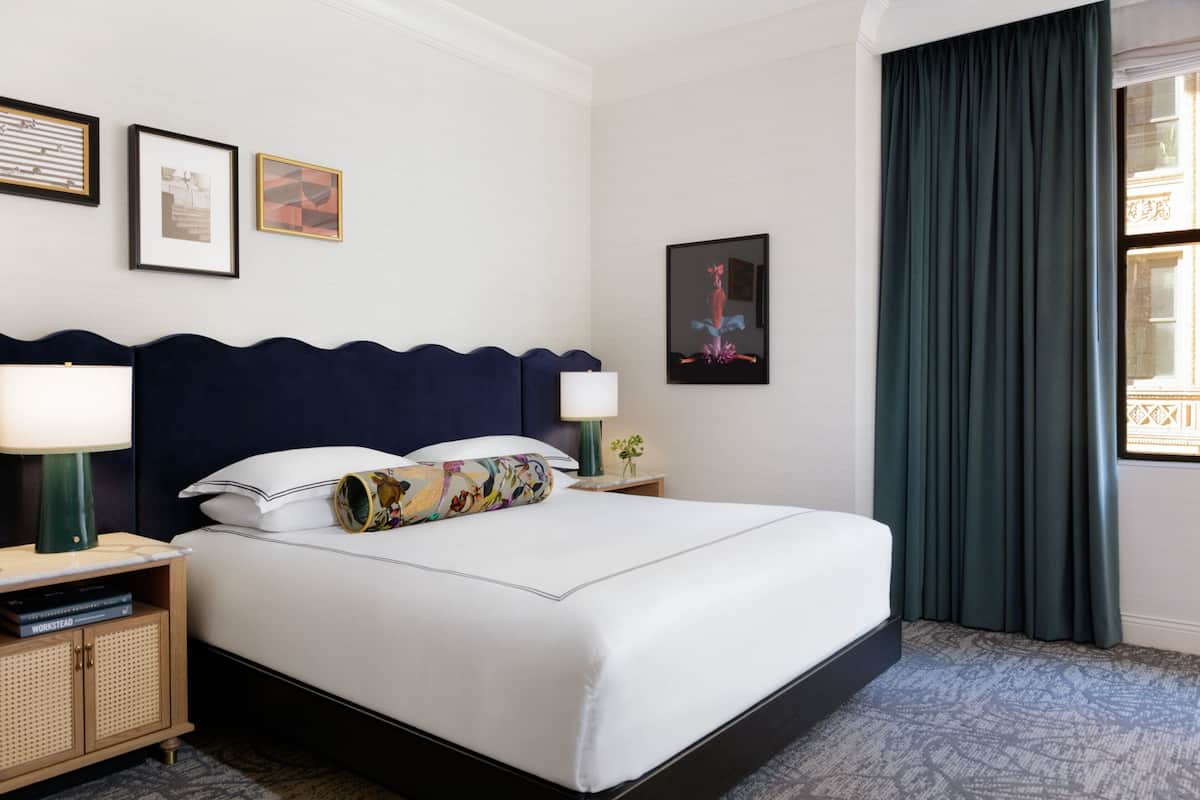 A hotel room with a large bed, two bedside tables, a nightstand, a framed picture, and a window with curtains.