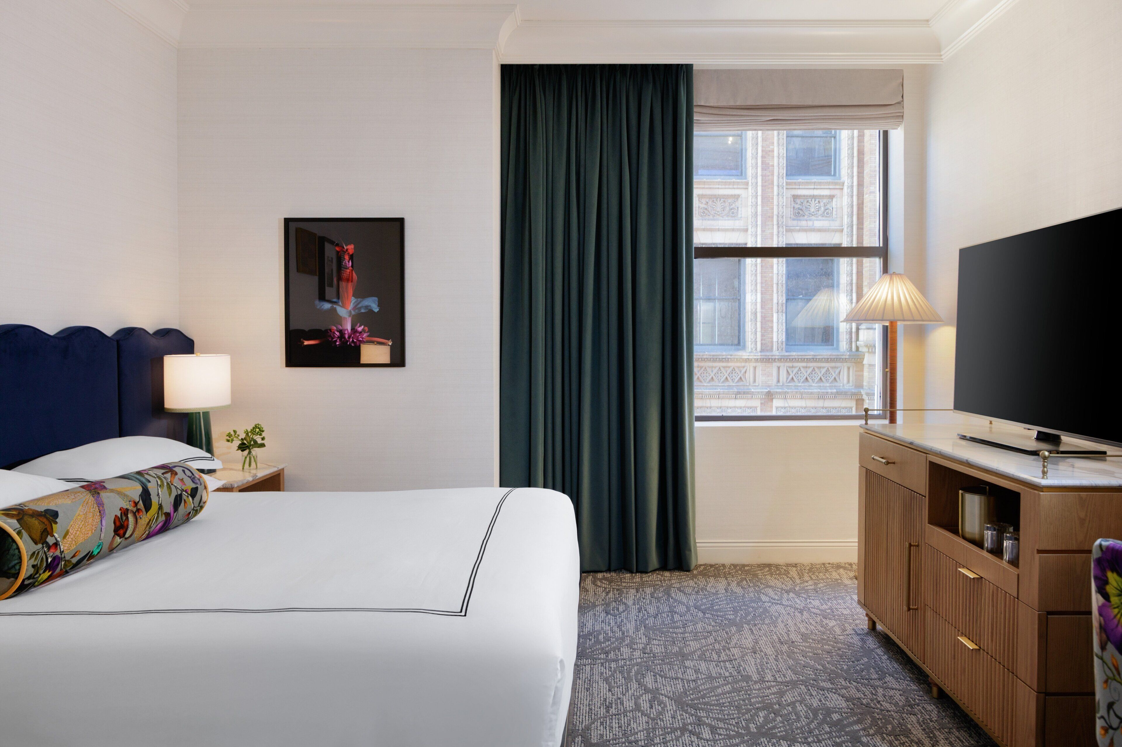 A hotel room with a bed, a TV, a desk, a lamp, a window with curtains, and a framed picture on the wall.