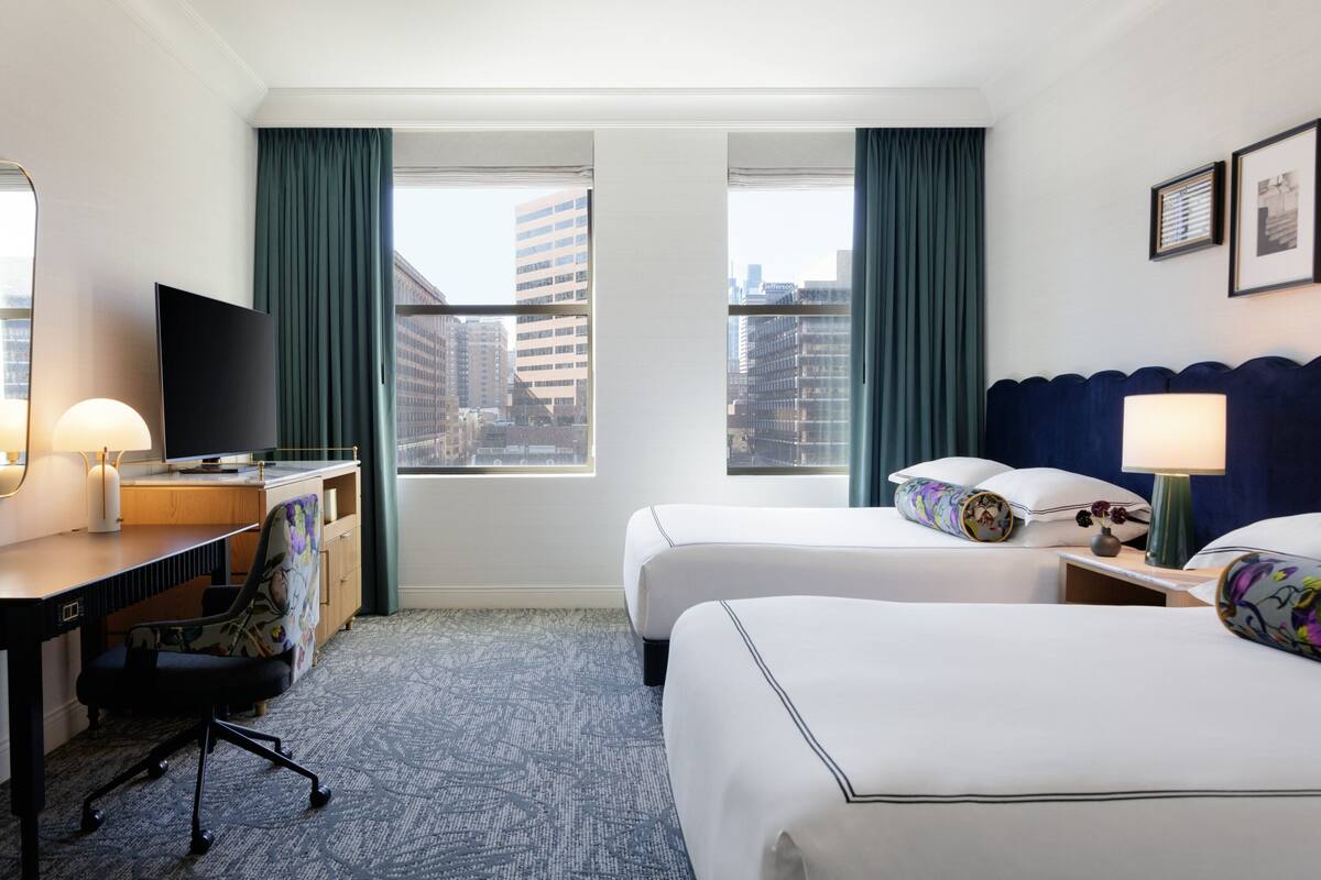 A hotel room with a desk, two beds, a TV, and a city view.