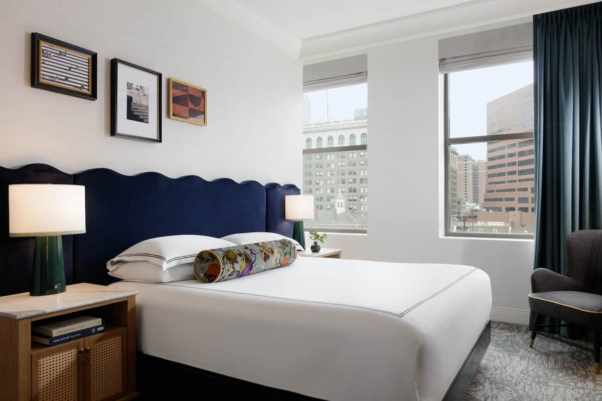 A hotel room with a large bed, two bedside lamps, a nightstand, a chair, and a window with a city view.