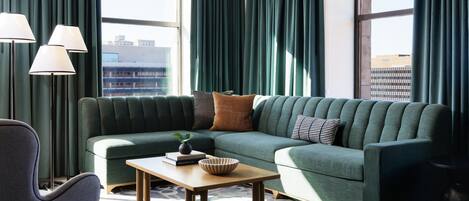 A modern living room with a green sofa, a wooden coffee table, and a view of the cityscape through large windows.