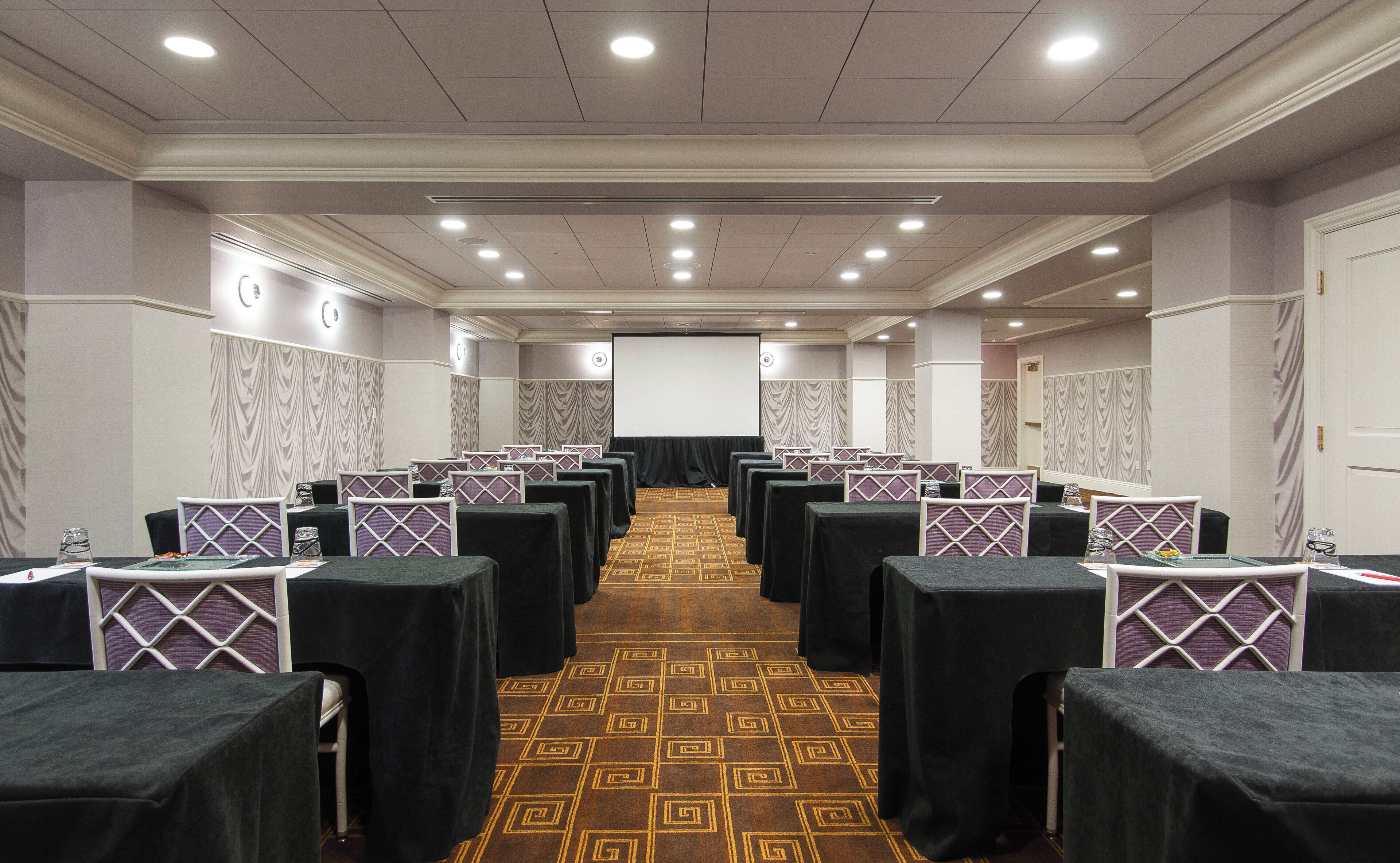A conference room with rows of tables and chairs, a projector screen, and a patterned carpet.