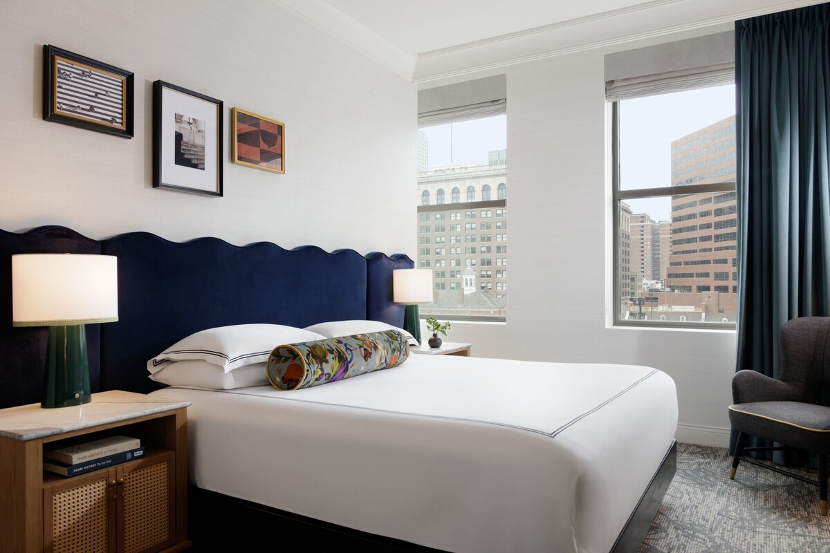 A hotel room with a large bed, two bedside lamps, a nightstand, a chair, and a window with a city view.