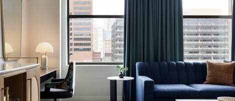 Ein modernes Hotelzimmer mit großem Fenster, einem blauen Sofa, einem kleinen runden Tisch, einem Schreibtisch mit Lampe und einem Spiegel.