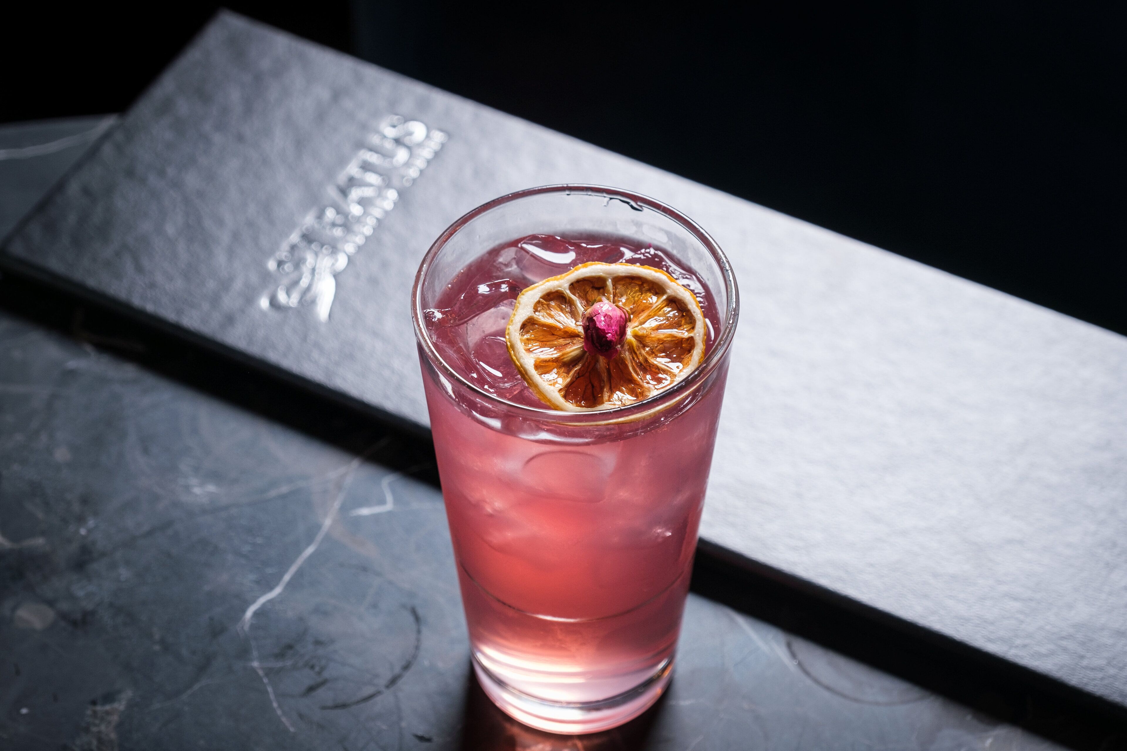A glass of pink beverage with a dried orange slice garnish on a dark surface.
