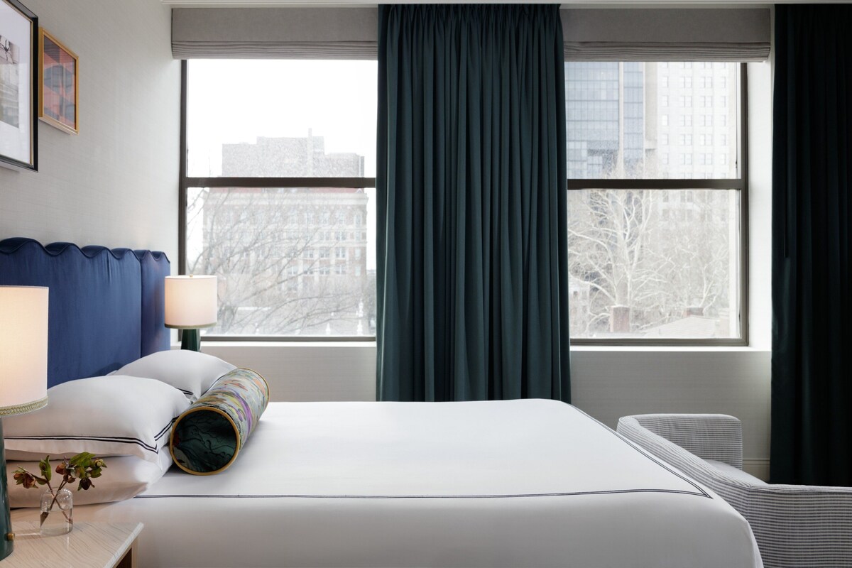A hotel room with a bed, a nightstand with a lamp, a window with curtains, and a radiator.