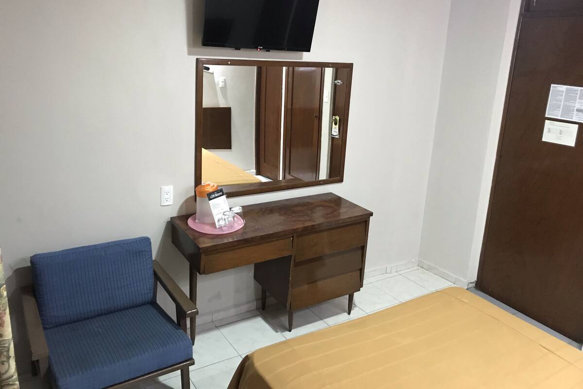 Habitació amb un llit, una cadira, un taulell de fusta, un mirall, una televisió penjada a la paret i una porta.