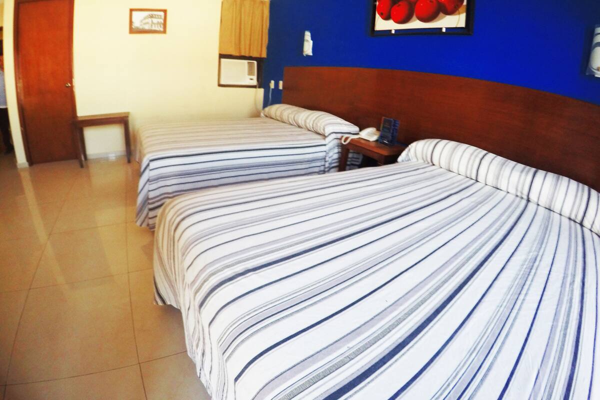 Habitació d'hotel amb dos llits, un panell de fusta al capçal, una taula petita i un aire condicionat fixat a la paret.