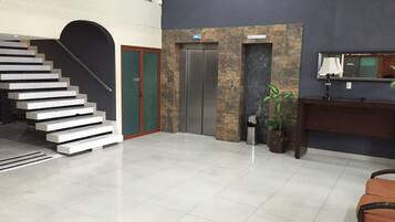 A modern lobby with a staircase, an elevator, a reception desk, and a potted plant.