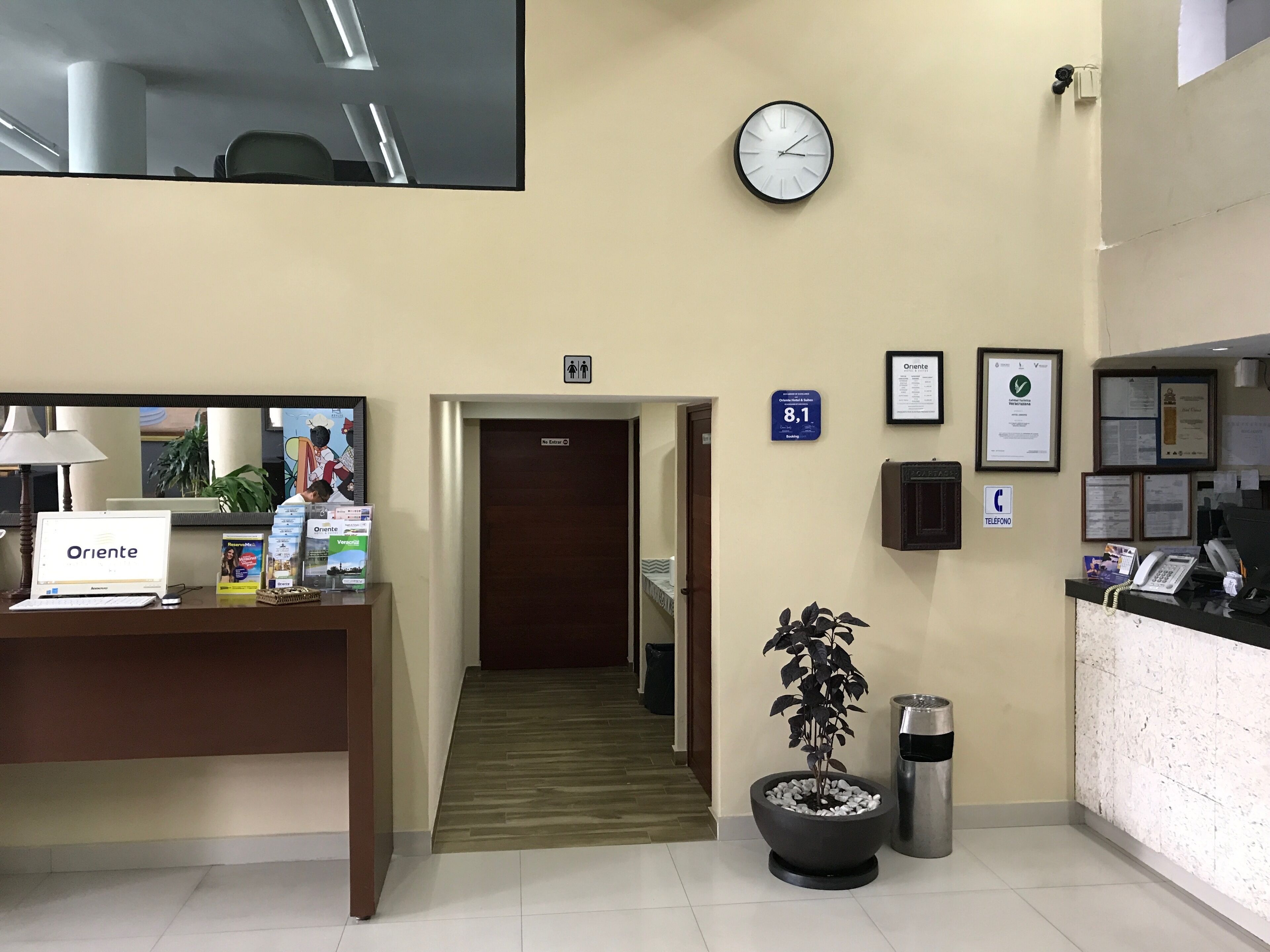 Zona de recepción con mostrador, computadora y carteles informativos. Se observan también un reloj, un bote de basura y una planta en maceta.
