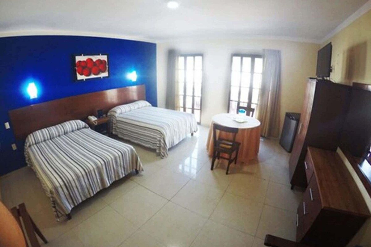 A hotel room with two beds, a dining table, a TV, and a large window.