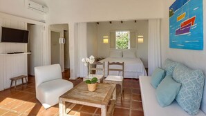 Une pièce avec un canapé blanc, une table basse en bois ornée d’une plante, un téléviseur fixé au mur et un lit à baldaquin blanc.