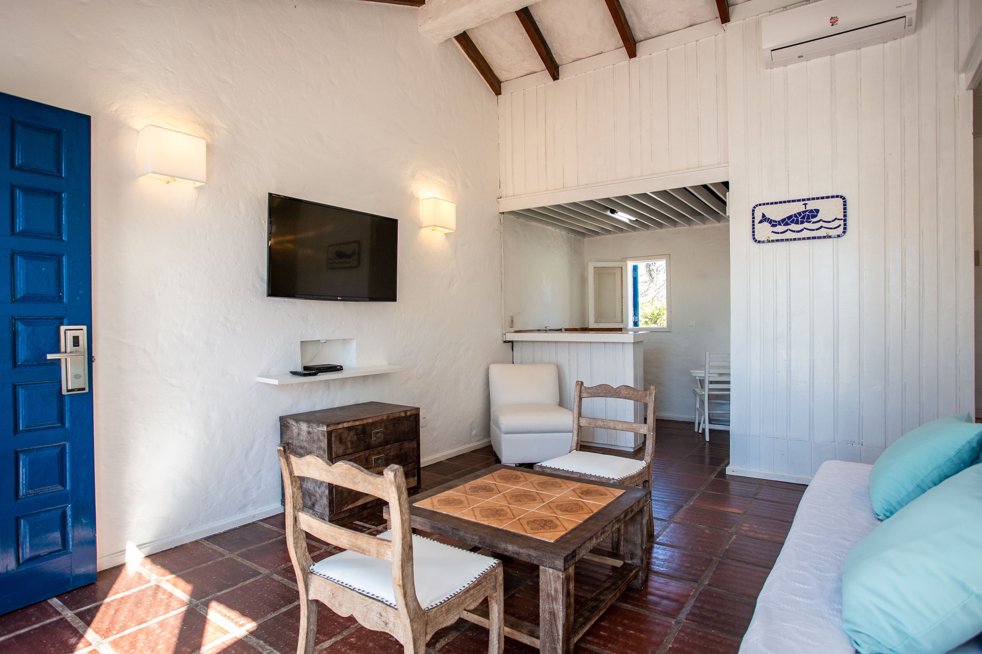 A room with a blue door, a TV mounted on the wall, a wooden table with chairs, a white sofa, and a tiled floor.
