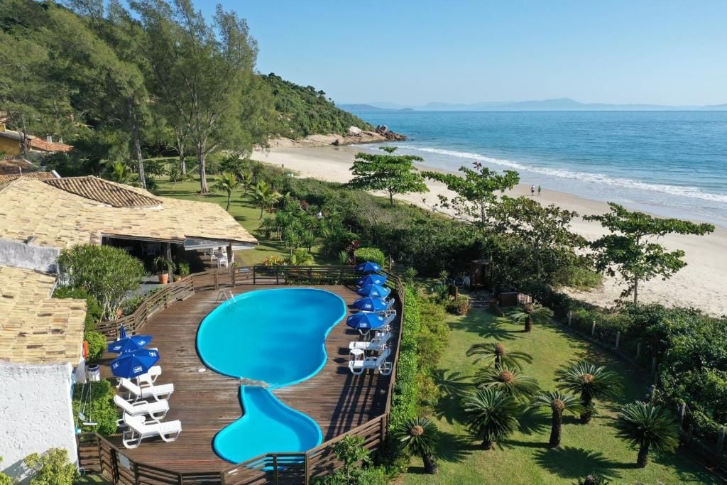 A swimming pool with a wooden deck, surrounded by lounge chairs and umbrellas, overlooking a beach and ocean.