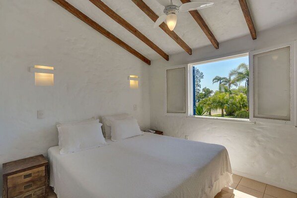 A bedroom with a bed, wooden beams, a ceiling fan, and a window with a view of palm trees.