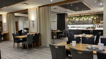 Um restaurante moderno com mesas e cadeiras de madeira, um espaço de bar e uma cozinha visível através de uma divisória de vidro.