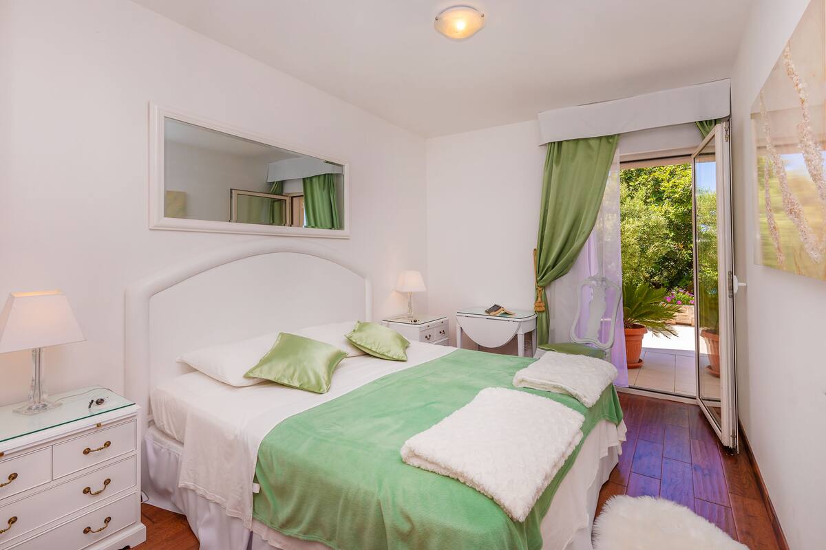 Un dormitorio con cama, mesitas de noche, un espejo y vistas a zonas verdes a través de un ventanal.