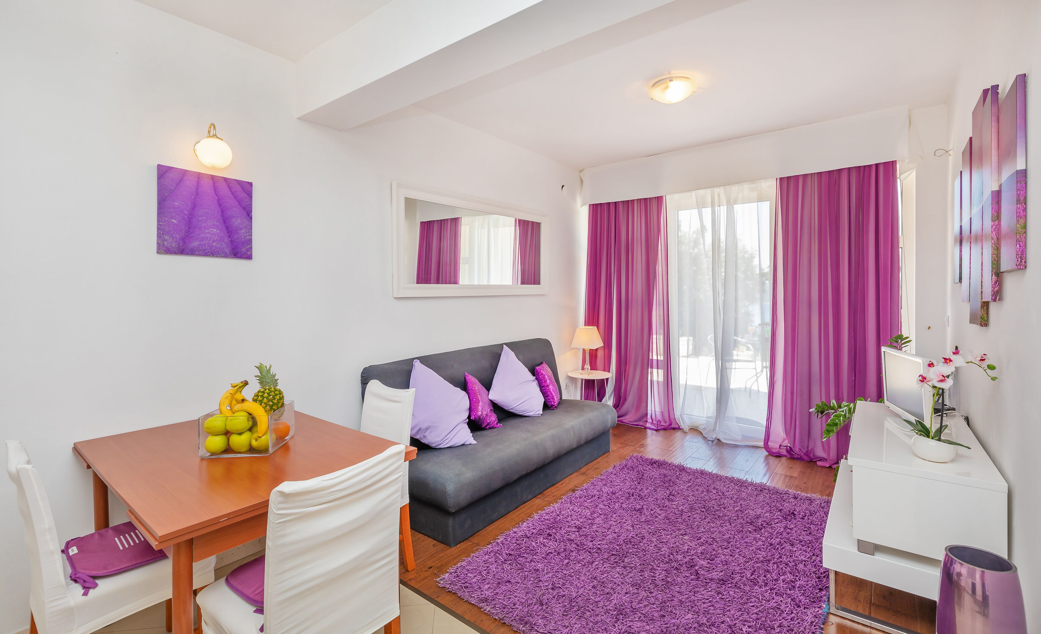 A modern living room with a grey sofa, purple cushions, a wooden dining table with fruit, and a purple rug.