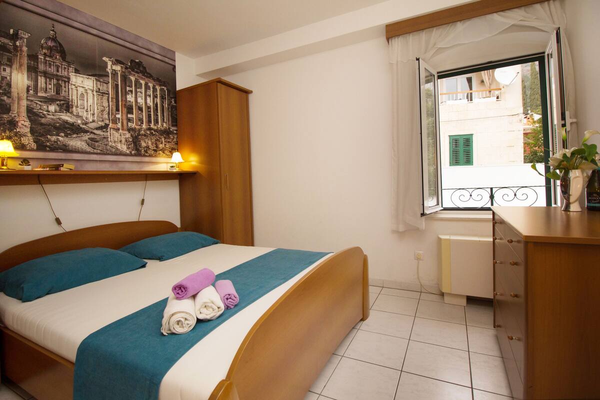 Cama bem arrumada com colcha azul-turquesa e branca, dois travesseiros e toalhas dobradas. Um guarda-roupa de madeira fica ao lado da cama, e uma janela com vista para a rua é visível.
