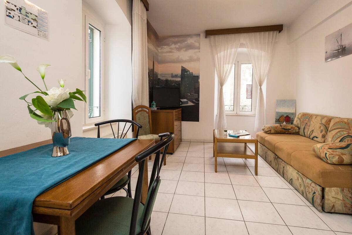 Sala de estar com uma mesa de jantar, cadeiras, sofá, televisão e um mural com vista para a cidade.